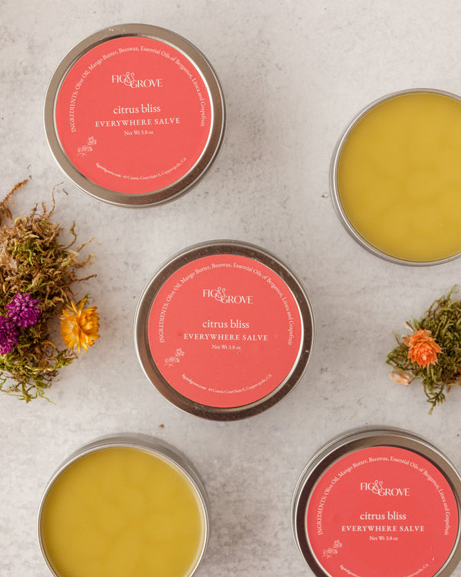Four tins of Citrus Bliss 'Everywhere Salve' with pink labels on a textured surface with flowers - Fig & Grove