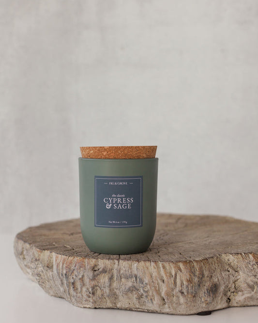 Cypress & Sage Candle in a sage green glass jar with cork lid on a wooden surface against a light gray background