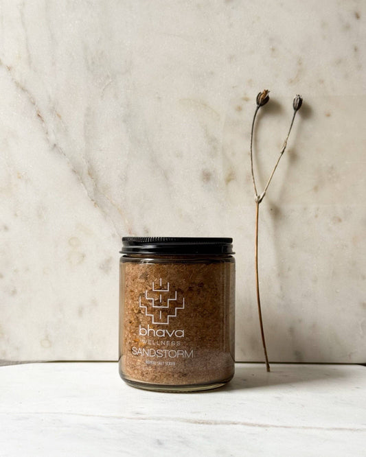 Jar of 'Sandstorm' candle by bhava on a marble surface with a dried plant.