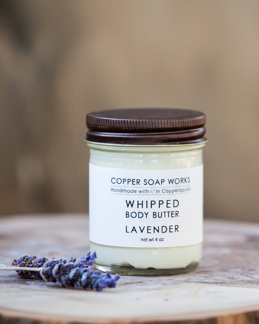 A jar of Whipped Body Butter with a brown lid, labeled 'Copper Soap Works - Whipped Body Butter - Lavender'. The jar is placed on a wooden surface with a sprig of lavender beside it.