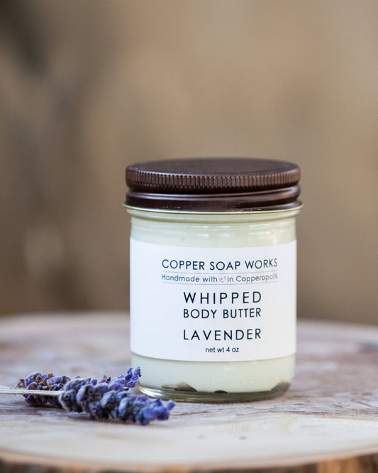 A jar of Whipped Body Butter with a brown lid, labeled 'Copper Soap Works - Whipped Body Butter - Lavender'. The jar is placed on a wooden surface with a sprig of lavender beside it.